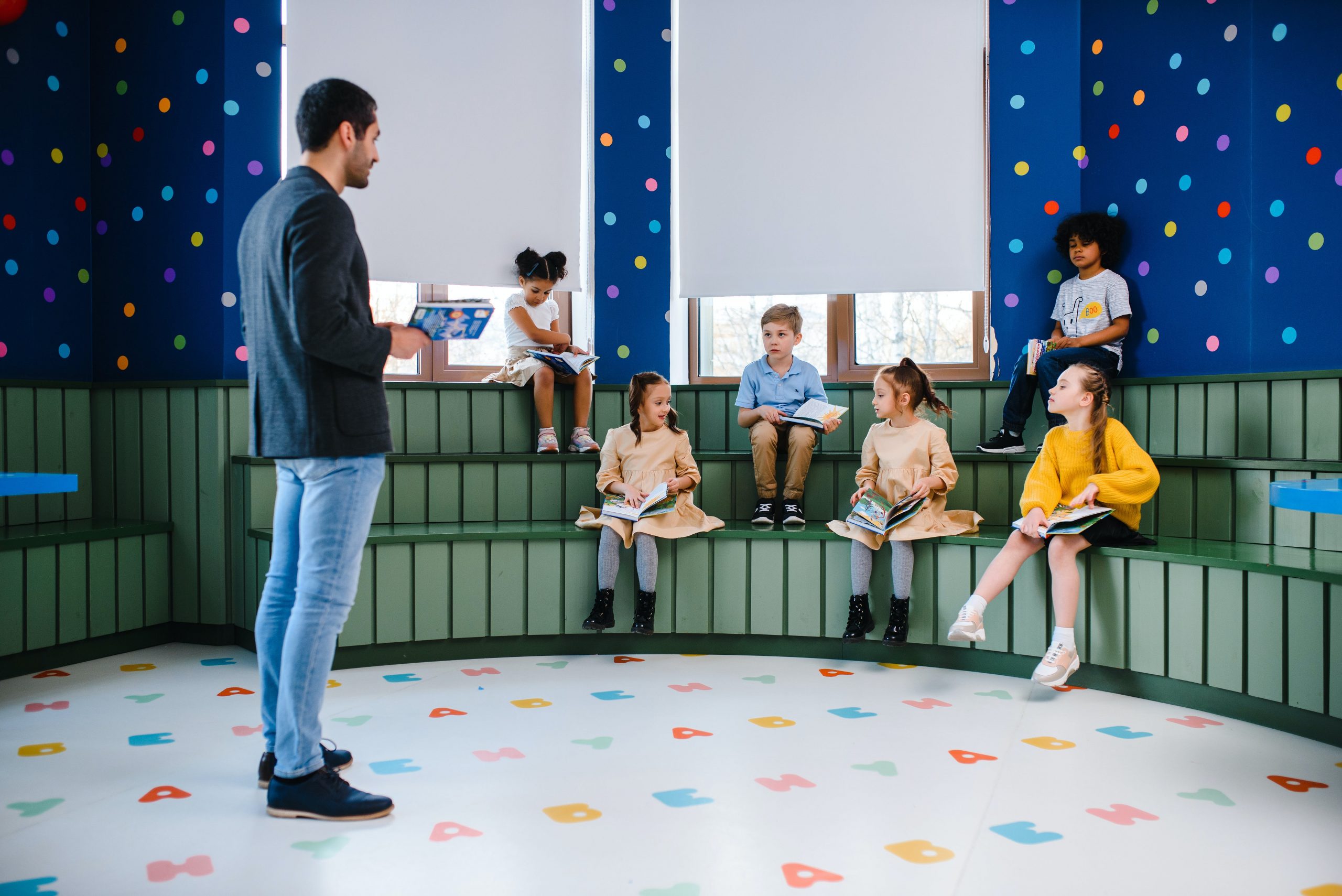 Teacher reading to attentive children sitting on steps in a colorful, modern classroom. 