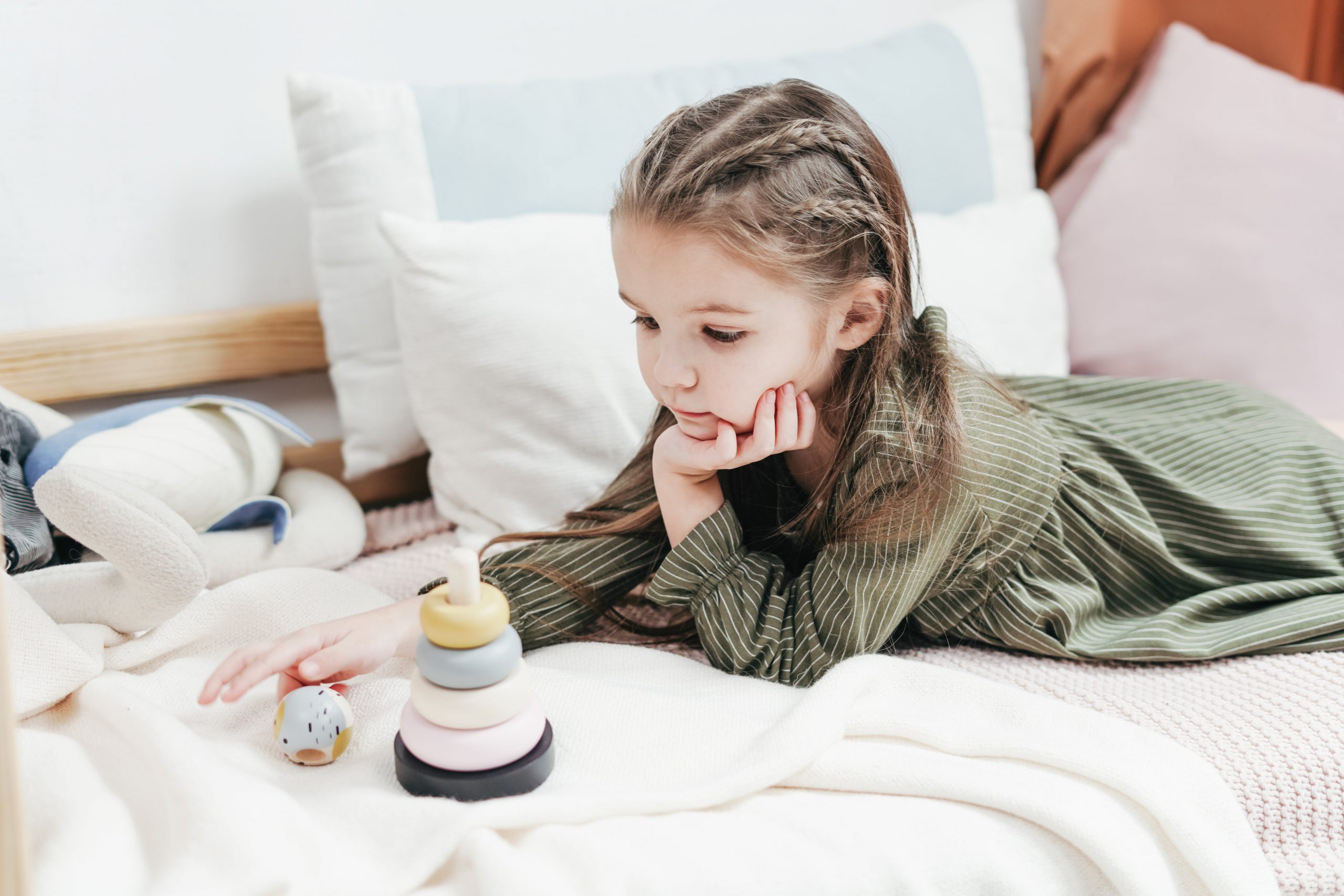A contemplative young girl lies on a bed playing with a wooden toy, surrounded by pillows. 