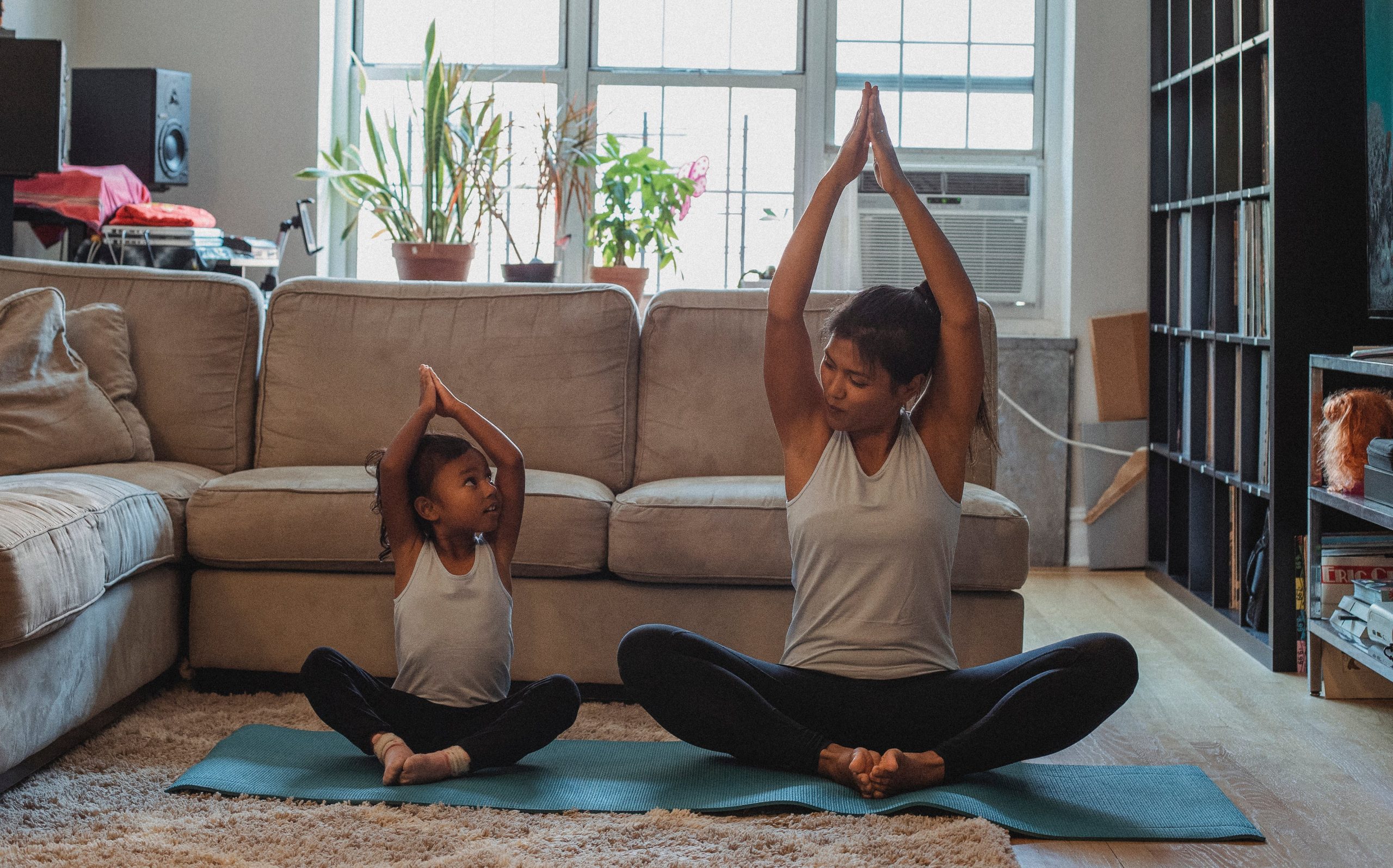 Adult and child practicing yoga together with hands clasped overhead in a bright, cluttered living room. 