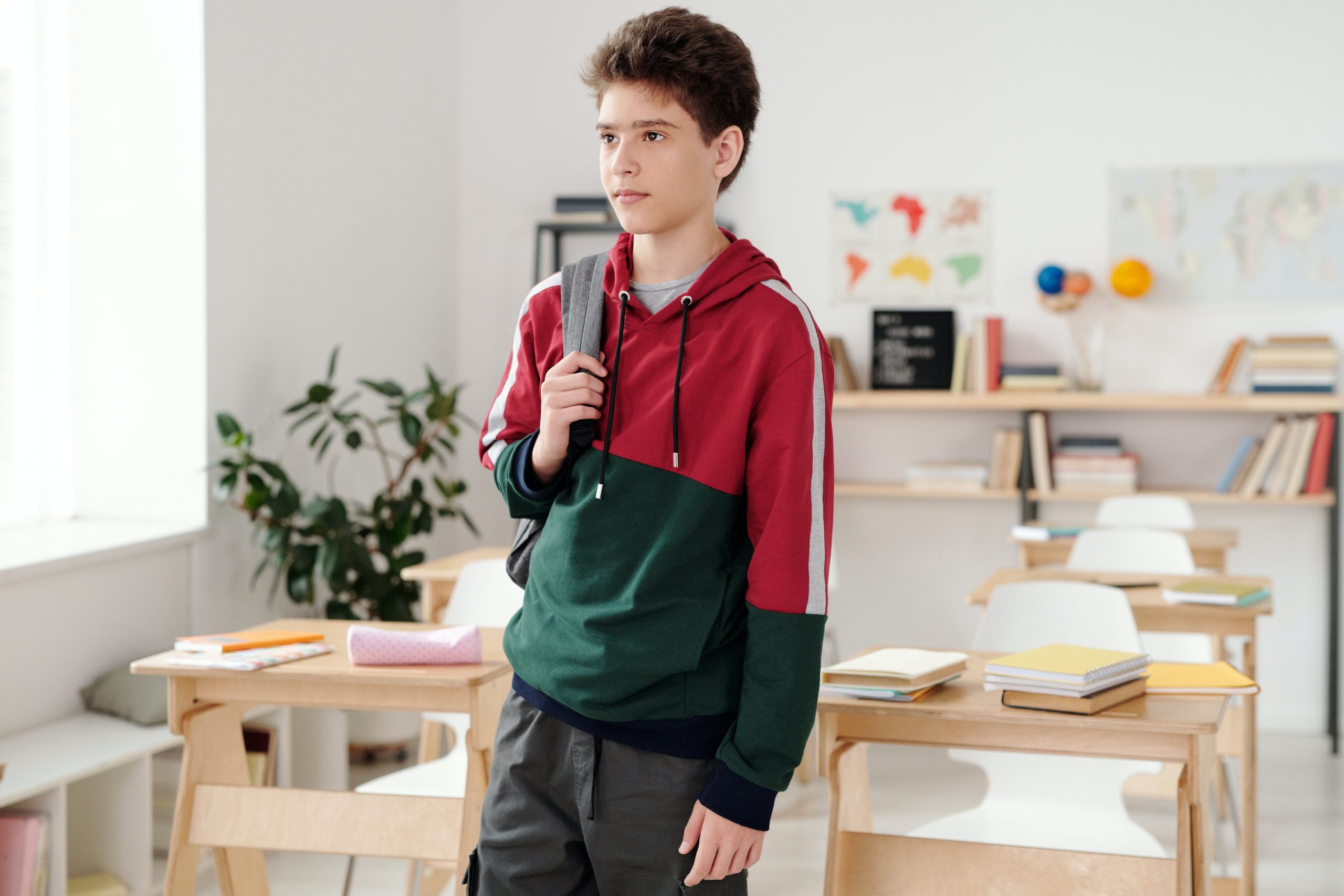 Teenage boy with backpack standing in a classroom with maps and books in the background. 