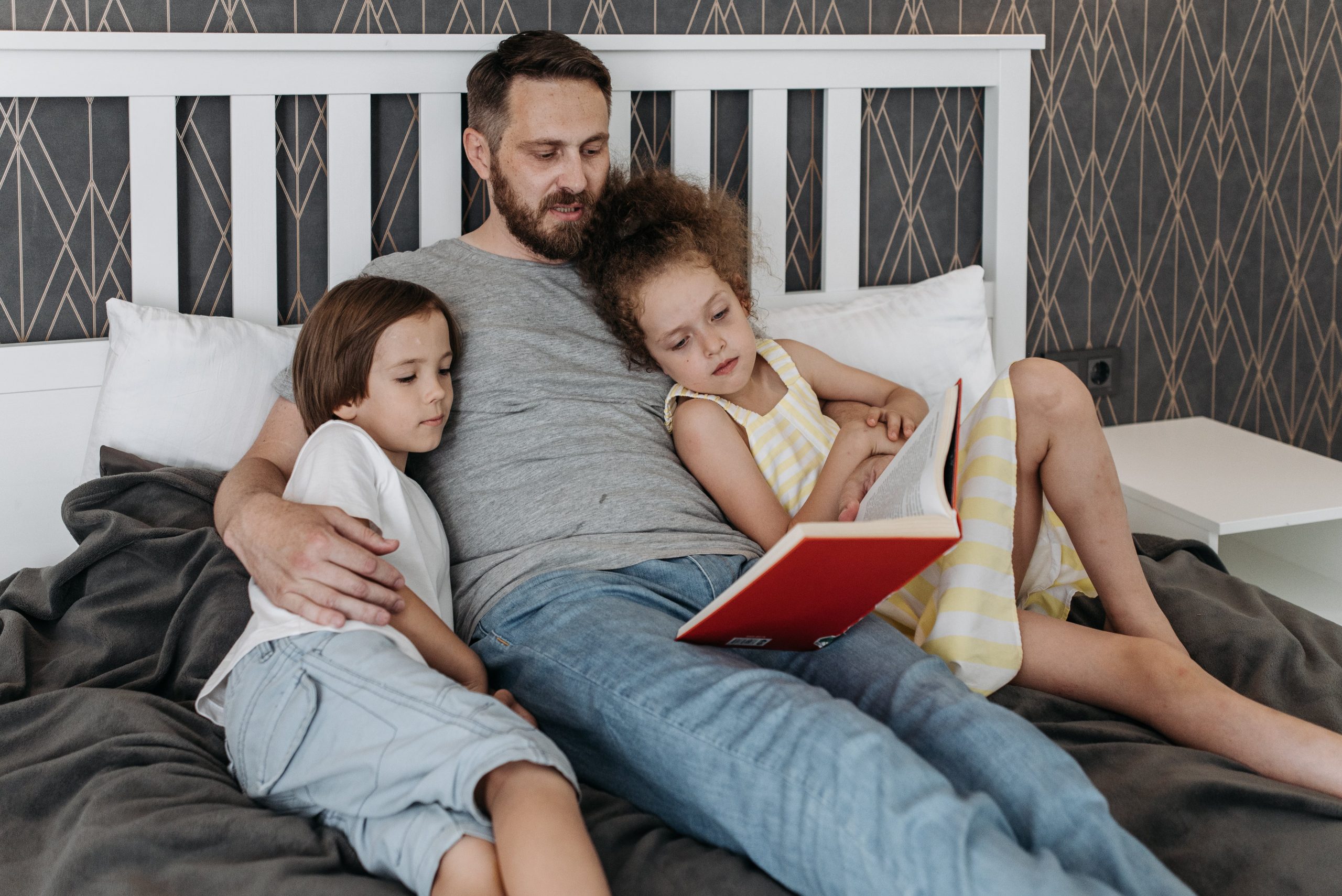 A father is reading a book with his son and daughter lying on the bed