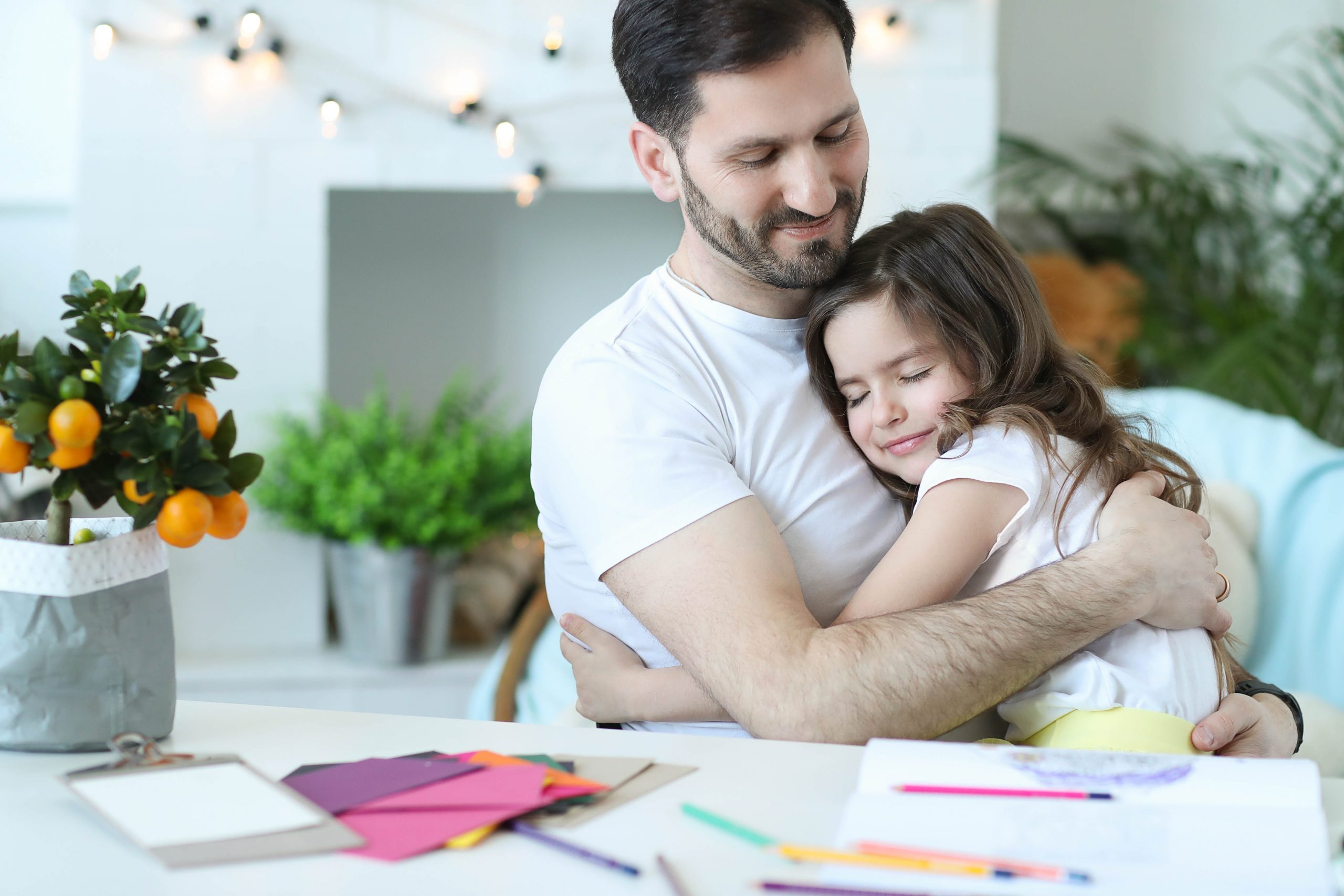 Affectionate father hugging young daughter, joyful expressions; creative art supplies scattered on table.