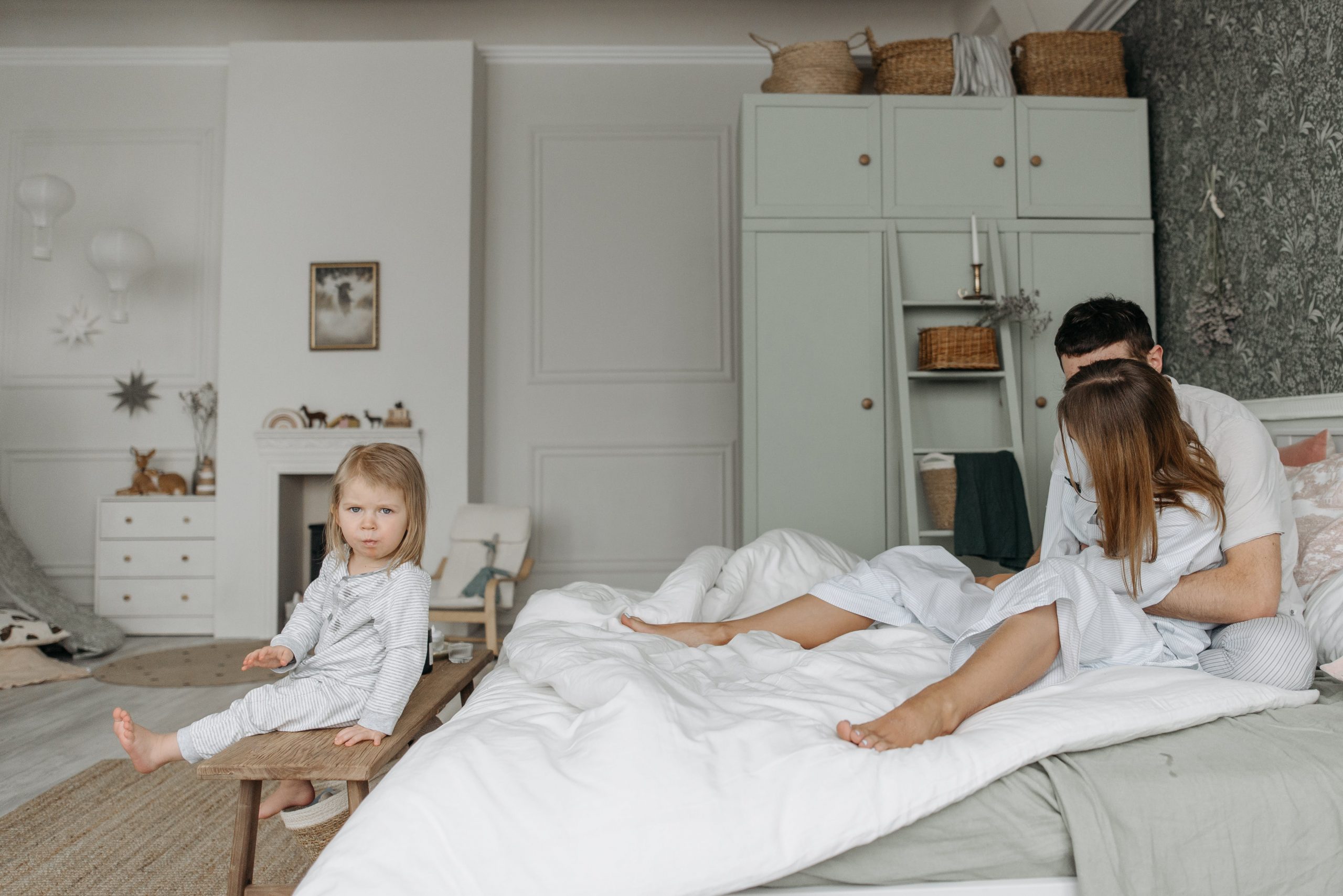 A toddler sits on a bench looking at parents cuddling in bed in a well-decorated room. 