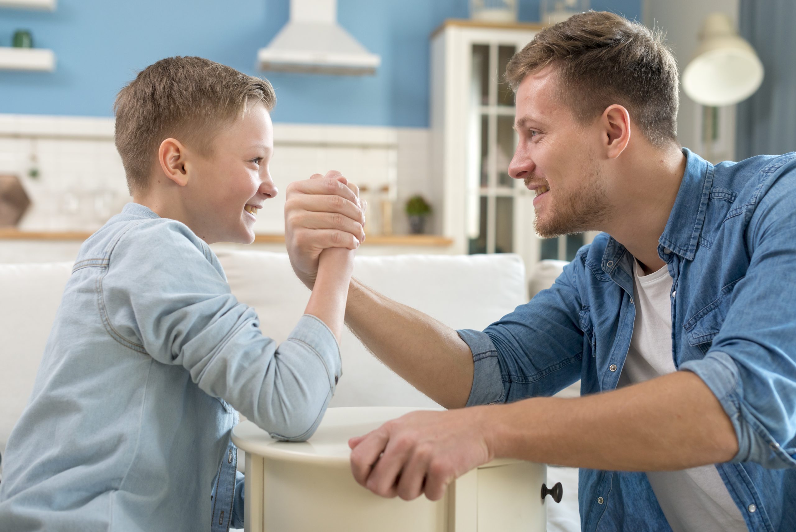 Father and son smiling and arm wrestling, bonding in a bright home setting. 