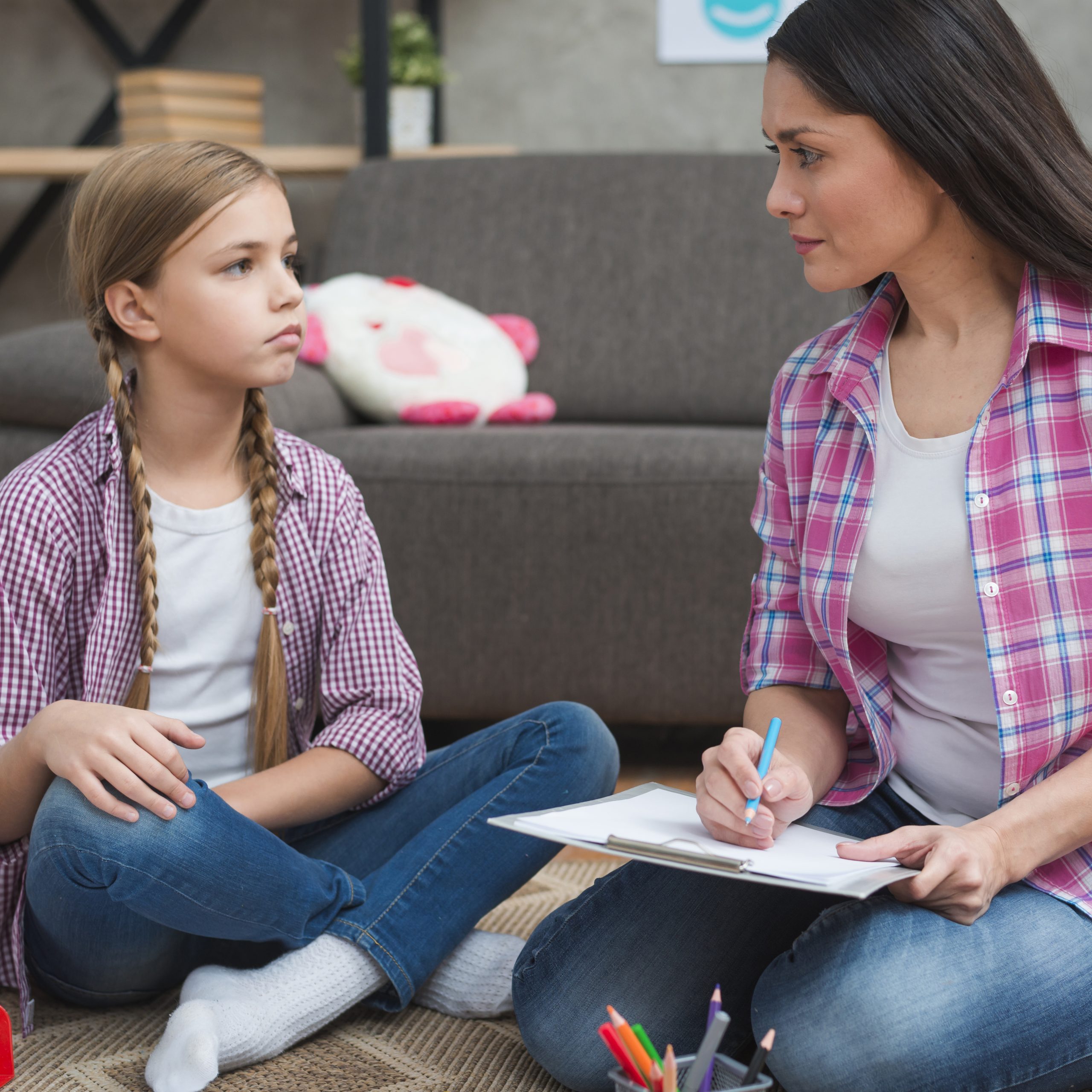 Child psychologist talking to a young girl, notepad in hand, in a cozy room setting.