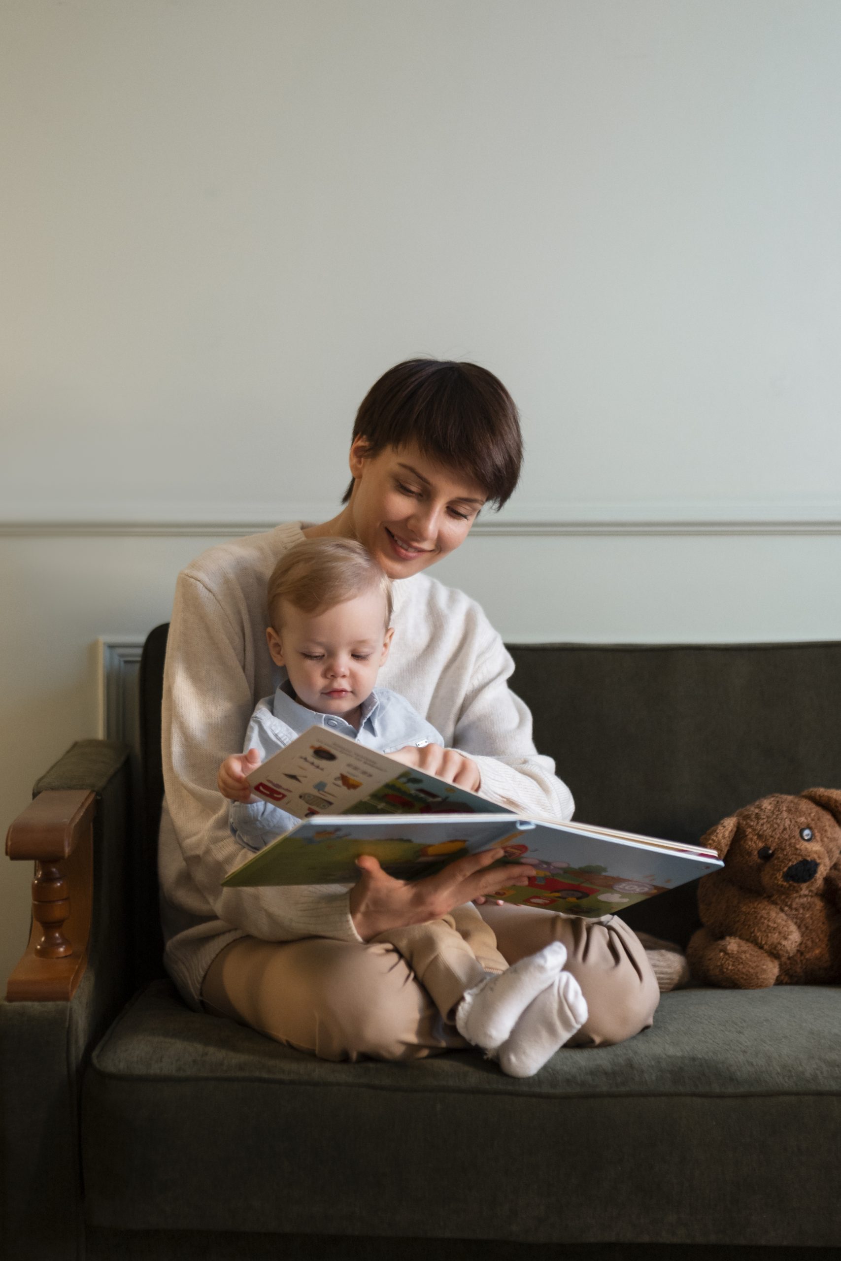 Mother and baby enjoying a book together on a couch with a teddy bear beside. 