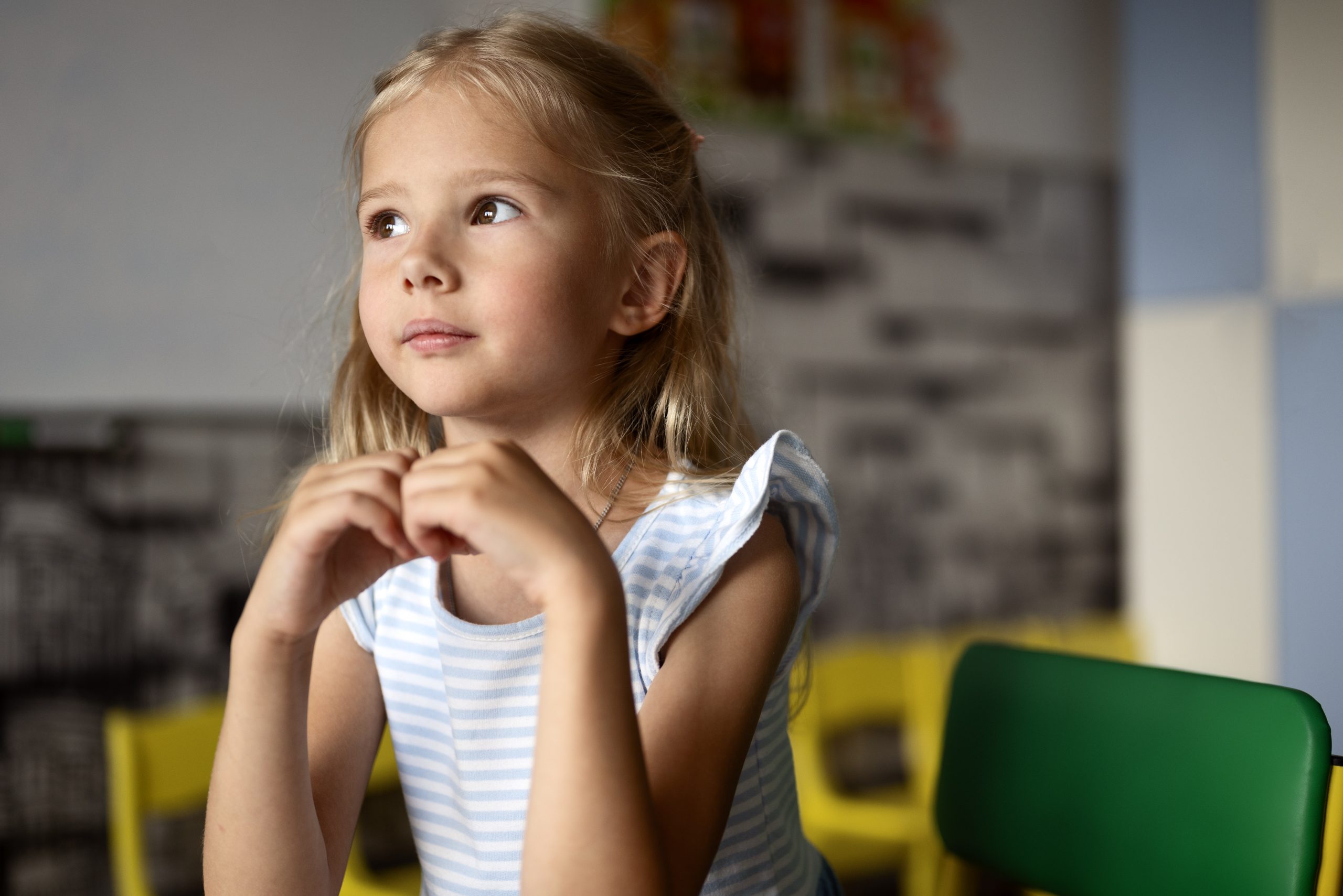 Young girl in striped dress sitting thoughtfully in a classroom with colorful chairs. 