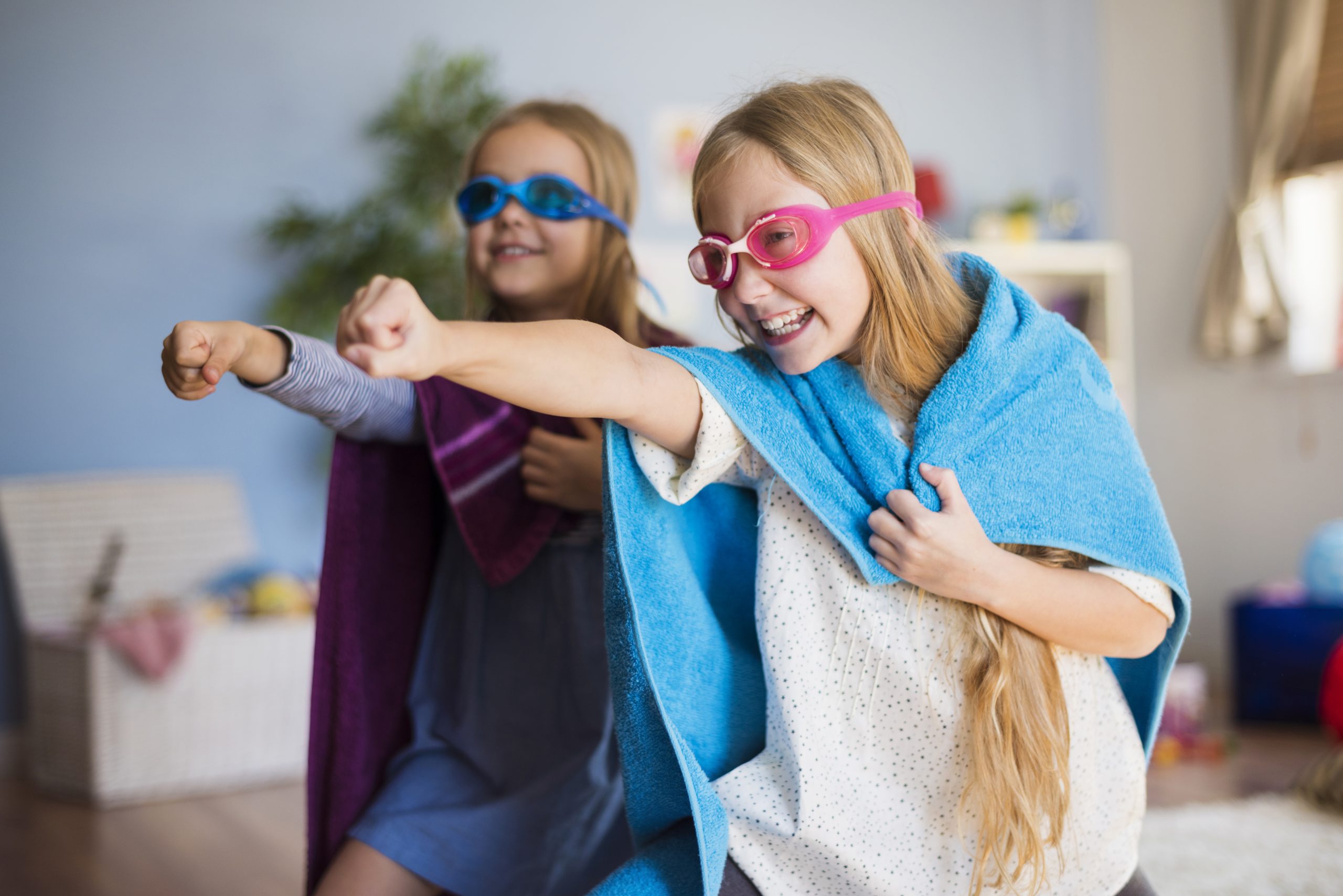 Two joyful children in goggles and capes play pretend as superheroes indoors. 