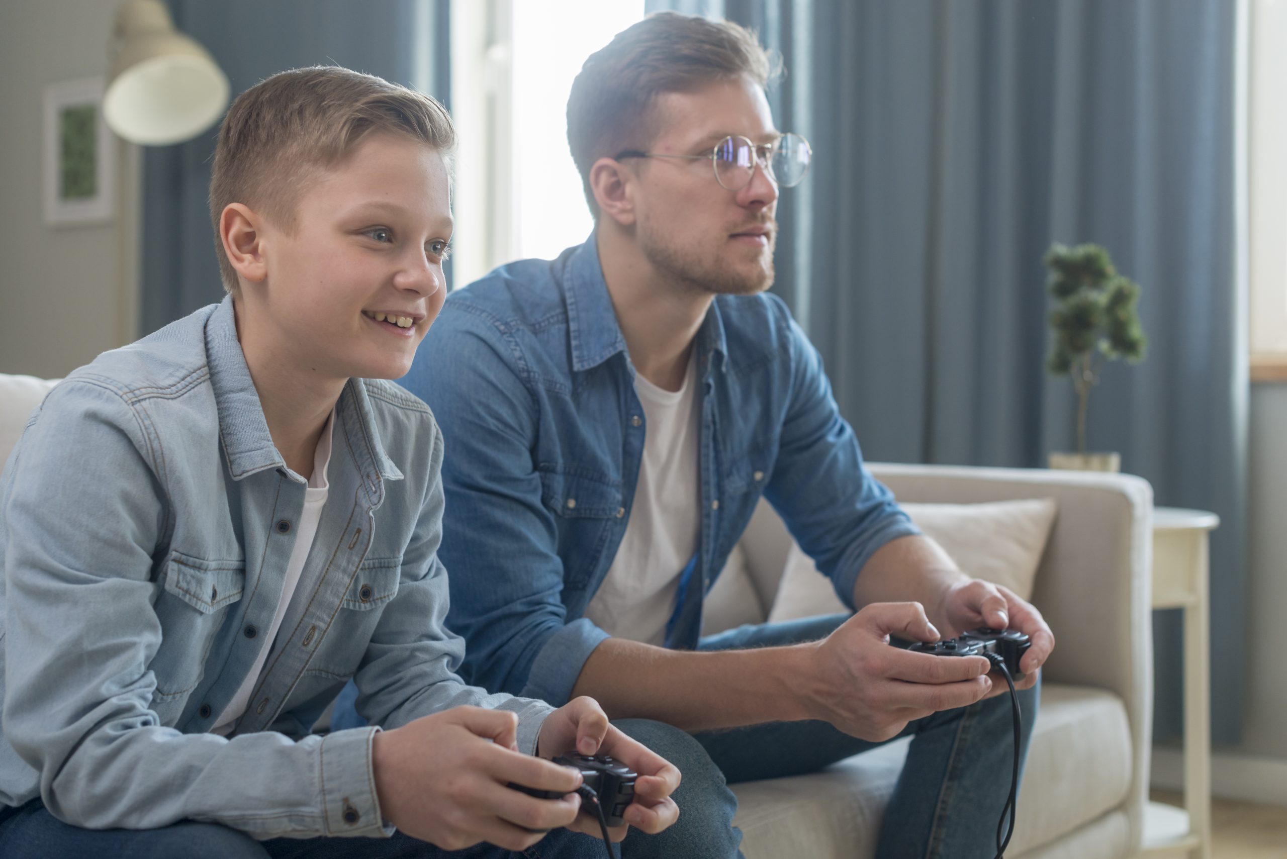 Smiling boy and focused man sitting on couch playing video games together in a living room. 