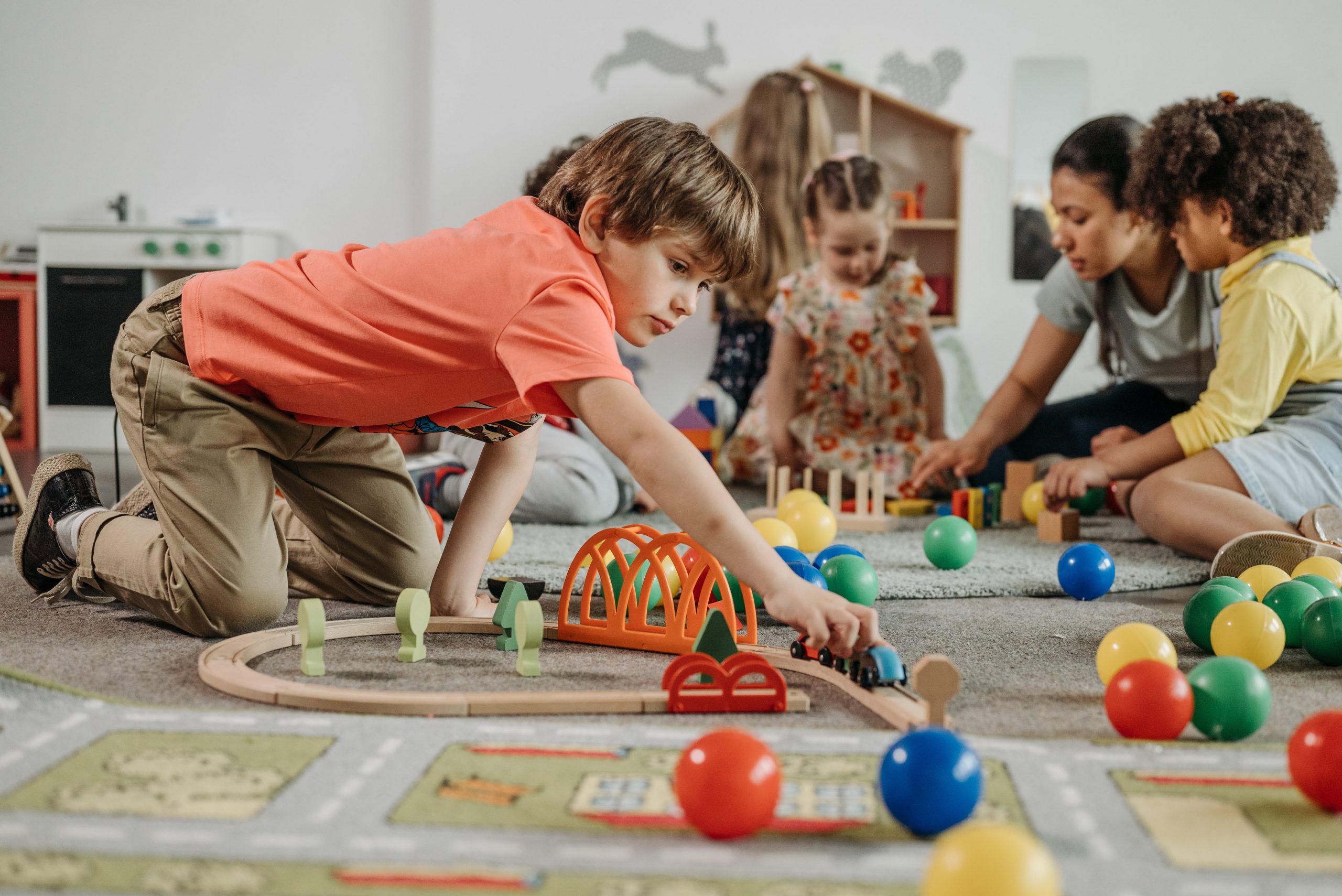 Children and a teacher play with a toy train and colorful balls on the floor in a classroom. 