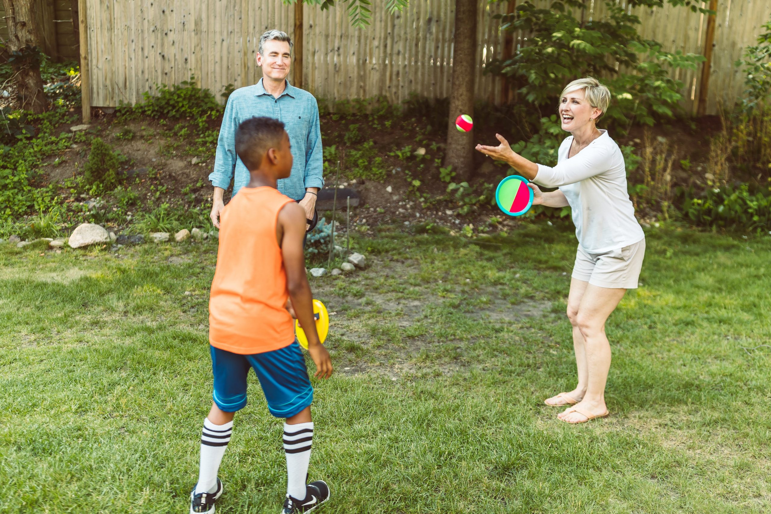 Family playing catch with a ball in a sunny backyard, with a man observing smilingly. 