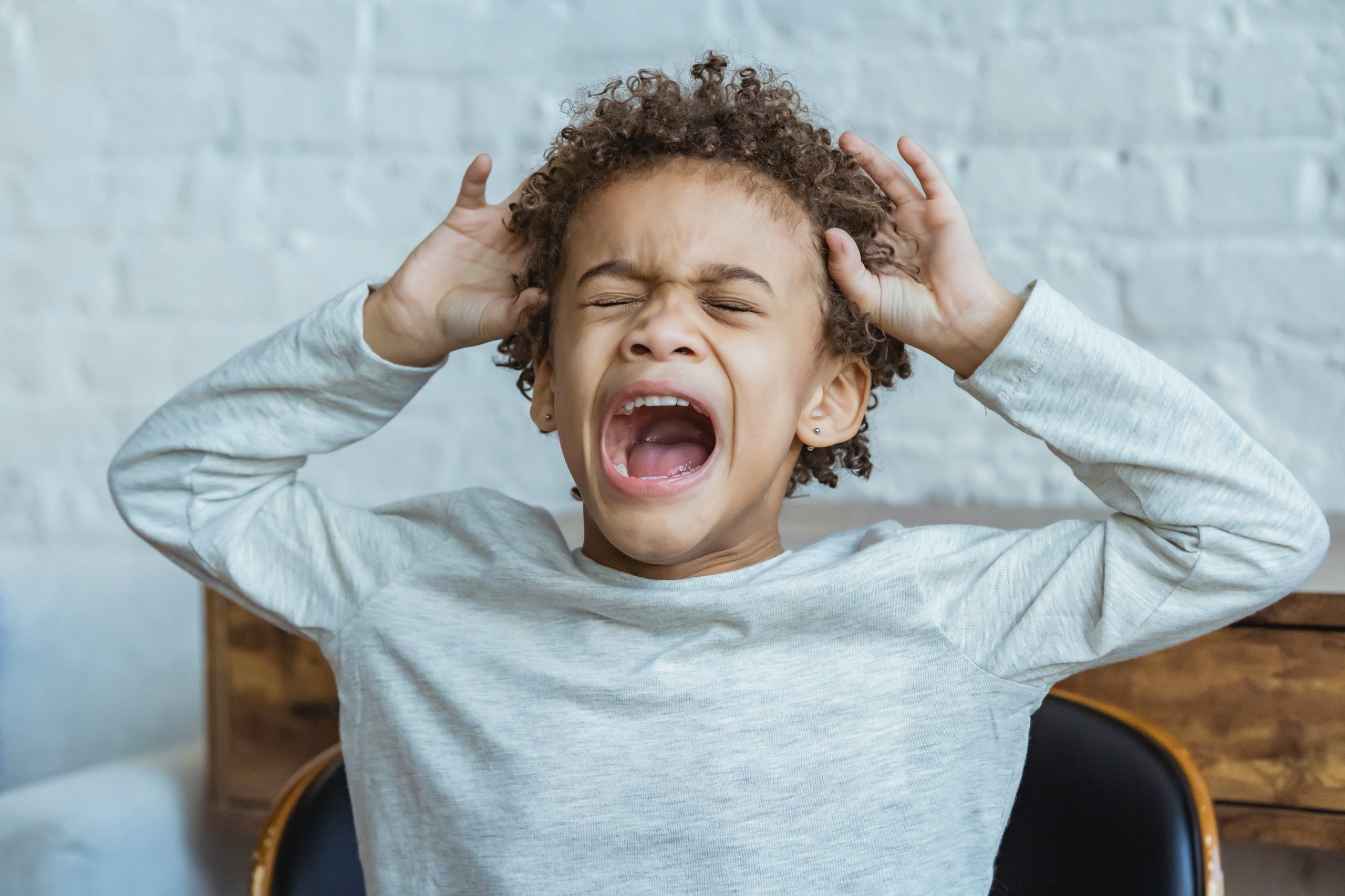 Frustrated child screaming, hands on head, standing against a blurred brick wall background. 