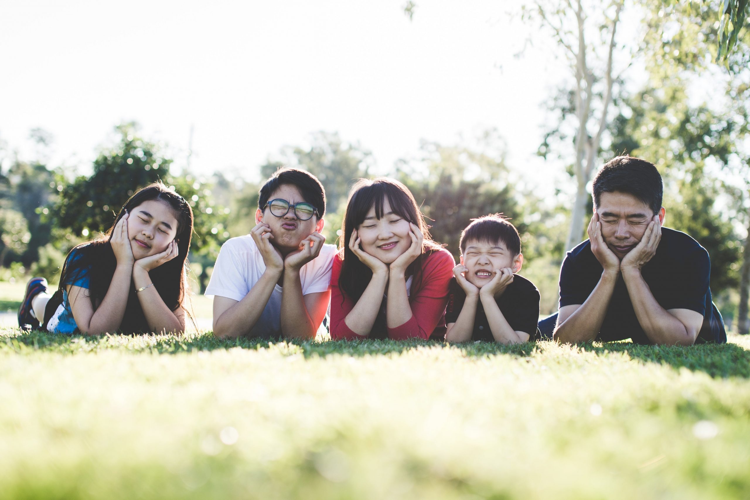 Family lying on grass in park, smiling with hands on cheeks, enjoying sunny day.