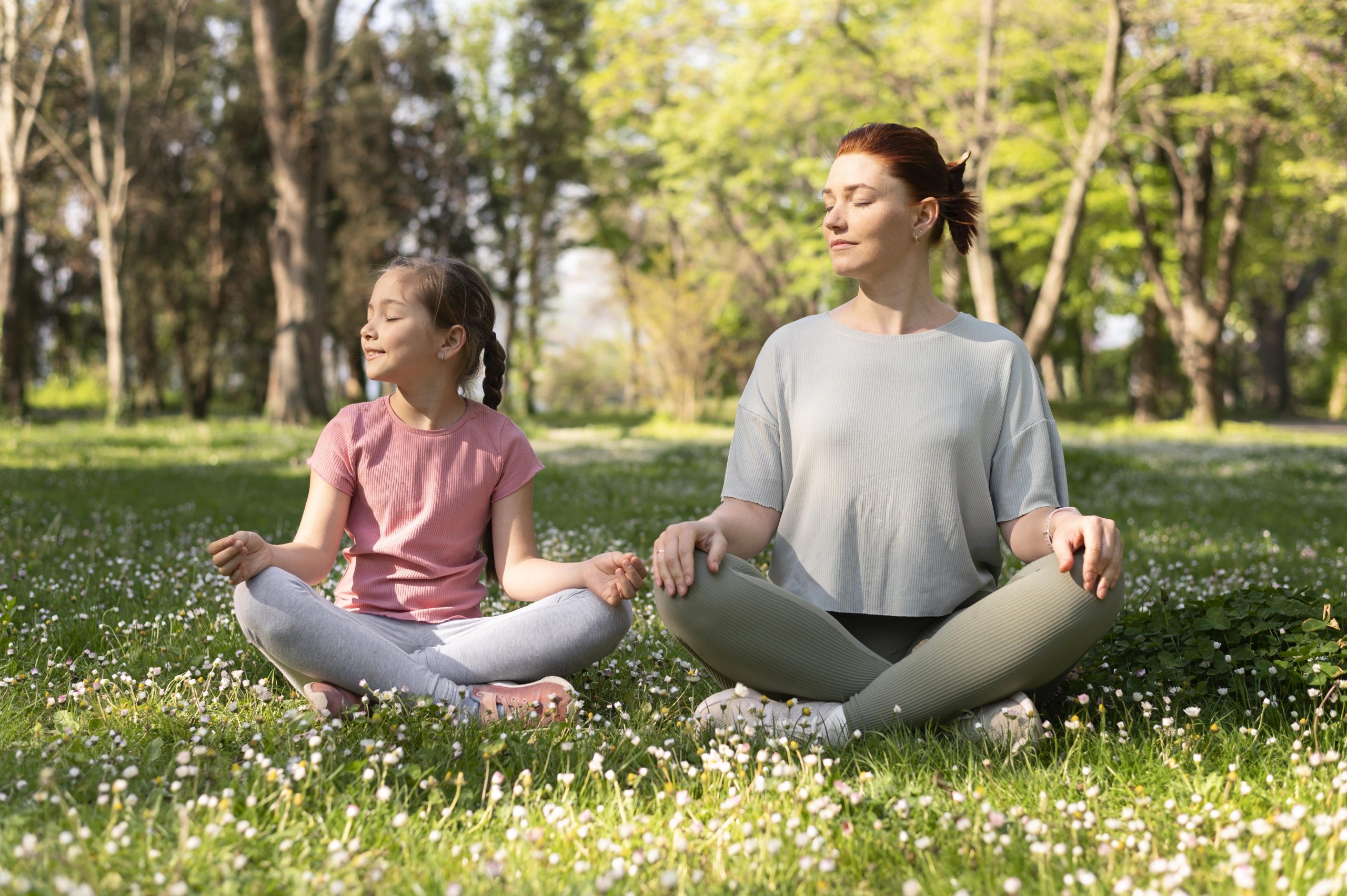 Woman and young girl practicing yoga together peacefully on a grassy field with trees. 