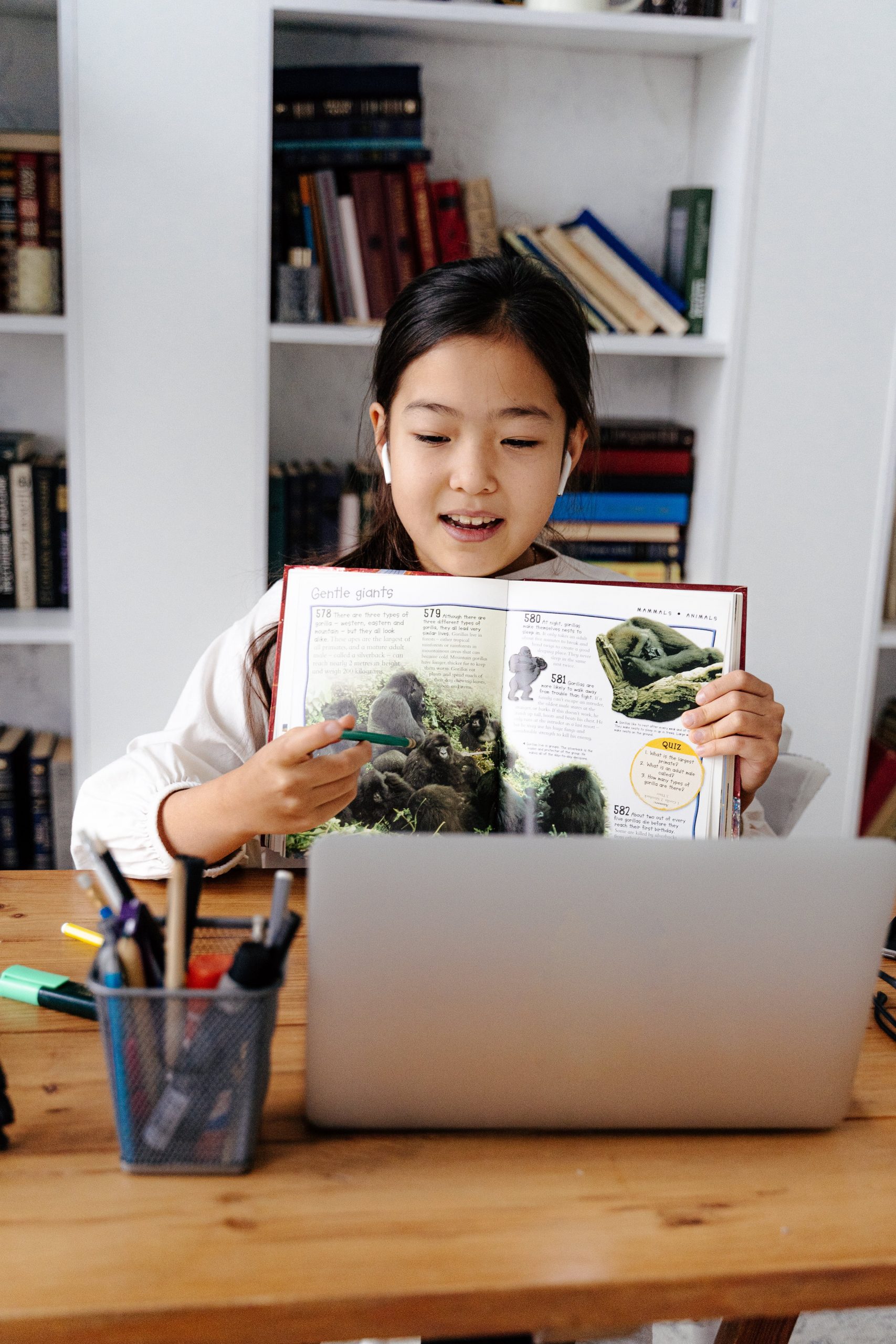Young girl reading a book aloud with earphones, sitting at a desk with a laptop. 